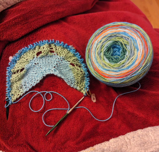 A small shawl start with a bit of lace knitting sits next to a ball of self-striping shawl yarn. The colours are heavy on the blues and greens but there's a lot of pops of other colours.