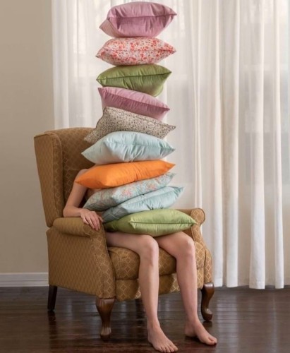 Photography. A color photo of a person sitting on a beige armchair with 10 small cushions on her lap. The (probably female) person with bare legs disappears behind this tower of different colored decorative cushions. Only a window with a white curtain can be seen in the background. 
Info: Brooke DiDonato is a visual artist from Ohio who attempts to depict the subconscious in a surreal way in her works. Compositions that blend the ordinary with the uncanny.
