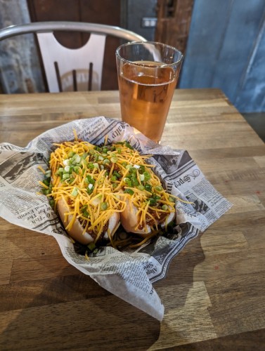 Chili dogs with cheese and green onions in a paper serving tray, and a glass of blackberry flavored hard cider.