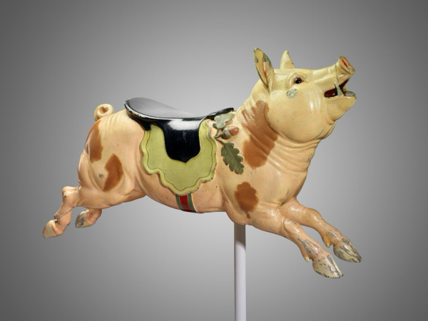 museum photo of the pig carousel figure on post in side profile on light grey background Pig is saddled and in running pose, light pink with brown spots, black and green saddle