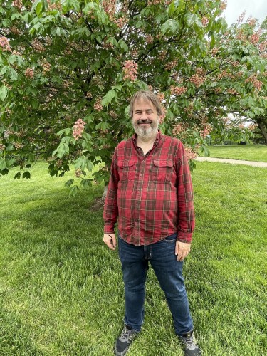 Chuck standing in front of a flowering tree. Red plaid shirt and jeans.