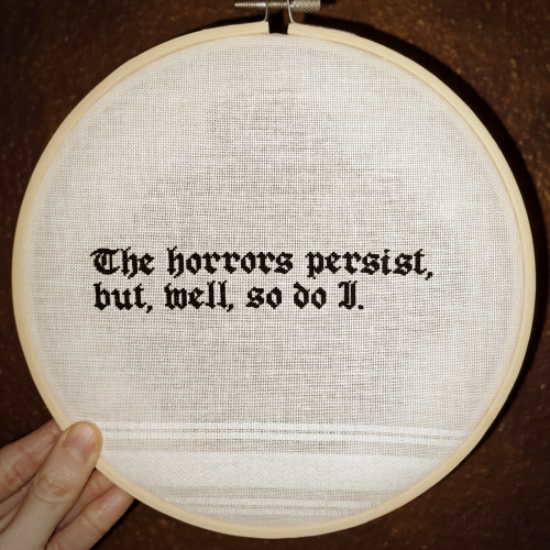 Cross stitch embroidery of the sentence "The horrors persist, but, well, so do I" in gothic type in black on white fabric in a hoop