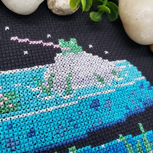 A close up of a cross stitch of a frog sitting on a rock trying to catch flies.