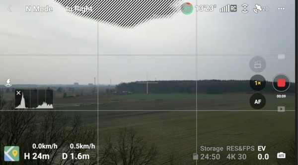 Widok z kamery drona na wiejski krajobraz z nałożonymi przezroczystymi elementami interfejsu wyświetlającymi status lotu, ustawienia kamery i elementy sterujące nagrywaniem.