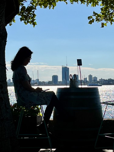 Profile of a woman sitting on a terrace waiting for her coffee; behind, the water of Matilda Bay
