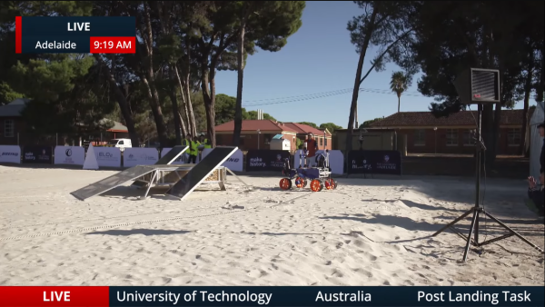 This is a photo of a robot competitor in the ARC.

The small dog-sized rover from Team QUT, has bright orange wheels and is currently driving on the sand around an obstacle.