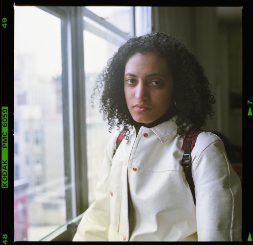 A woman with shoulder length, black, naturally curly hair and wearing a white leather jacket and backpack stands next to a sunny window. She gazes forthrightly, and maybe a little judgmentally, at the viewer.