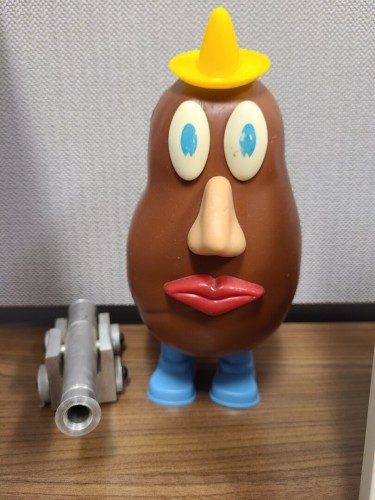 Photograph of an old, Mr. Potato Head doll standing next to a small, metal canon model on a wooden surface with a grey, fabric wall behind them. The Mr. Potato Head is a 1970s model with faded blue eyes, a yellow sombrero, dark red lips, light skinned nose, two tiny blue feet, and a dark brown body. The canon is small and silverish with four wheels, one of which is smaller than the rest.