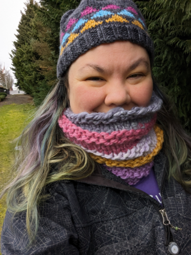Terri is standing in the backyard by the cypress trees in the cold. She is wearing a very thick handspun knit cowl with five colour stripes, and a knitted hat from her own pattern with triangles on it. 