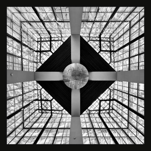 This image captures a striking architectural interior, presented in black and white, showcasing a complex glass ceiling intersected by a geometric metal framework. The central feature is a large, circular disk held by four symmetrical arms that split the view into quadrants. This disk is surrounded by a dark, diamond-shaped structure with textures that contrast with the transparent, patterned glass panels filled with visible steel supports. The ceiling structure forms a visually impactful pattern of lines and shapes, emphasizing the blend of transparency and solid elements against the light streaming through the glass.