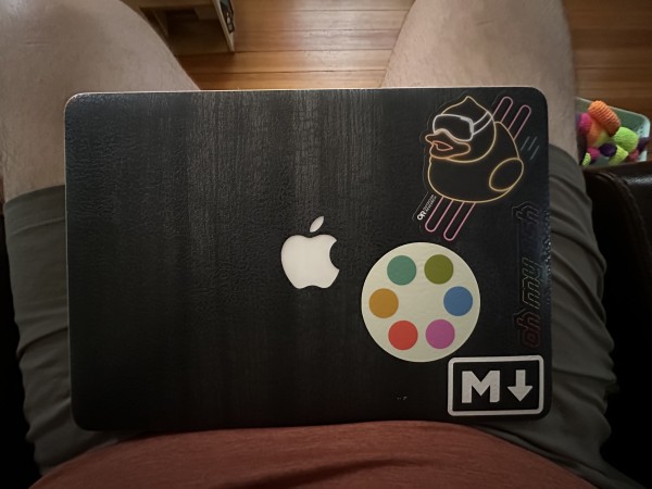 A top down shot of a MacBook of indeterminate specification that has a black charred wood skin applied to it. There are four stickers on the lid, one is a logo for the markdown formatting scheme, one is a black and rainbow Oh My ZSH logo, a neon rubber ducky sticker for Outdoor Research, and a Polymaths instance logo sticker with my favorite colors oriented to the top position.