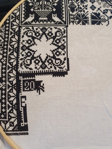 Black thread on white fabric, cross stitch. The number 20 can be seen. Above that is a large flower pattern. 