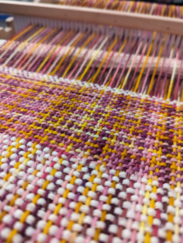 A weaving in progress on a rigid heddle loom. There are large stripes of white and a pink/yellow/purple variegated colour. 
