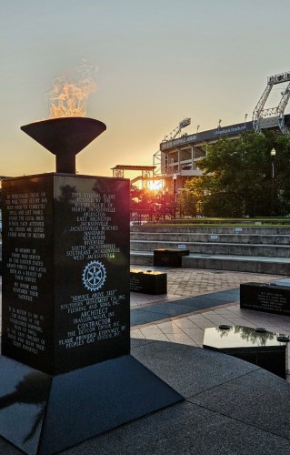 Standard next to a black marble pillar with an eternal flame coming from a bowl on top, etched with dedication to veterans of US military service. The view is at the large NFL stadium where a bright firey sun has peaked above the trees and illuminating the stadium and memorial beneath a clear blue sky.