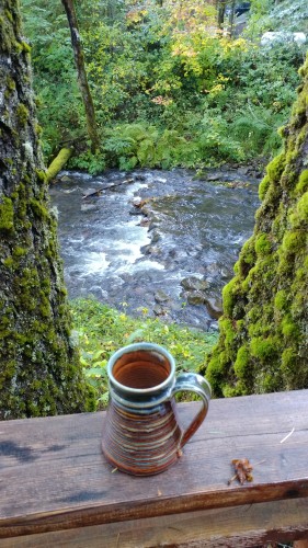 Coffee cup set on a railing looking between mossy trees at a creek