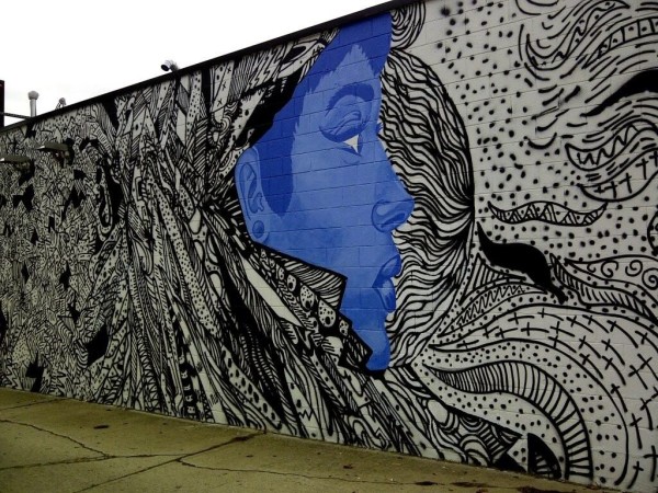 Graffiti artístico en un muro; el rostro de perfil de una mujer (en azul), entre figuras, líneas, motivos (en negro).