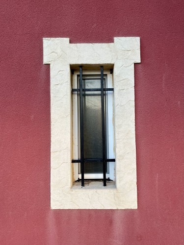 A narrow window with frosted glass and iron bars, set in a broad frame of white stone on a red wall