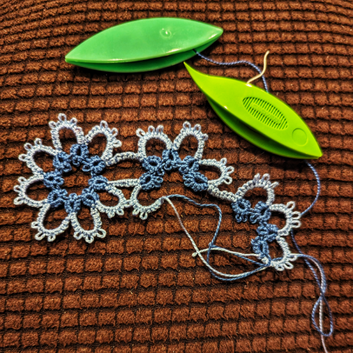 A tatted bookmark in progress sits next to two green tatting shuttles, only one of which is attached to the piece. The pattern is a set of three attached flowers and only one is complete with two half flowers dangling on one side 