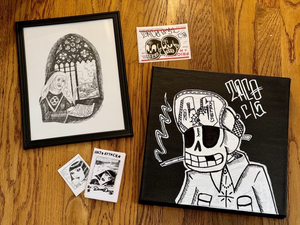 A photo of two pieces of art. Both are black and white and surrounded by stickers and mini comics. 
The piece of the left is a ball point pen drawing of a nun, near a stained glass window. It is drawn in Dame Darcy’s unique style. 
The other piece is a painting by Taco CS. It has his name written in a chino graffiti style and shows a skeleton wearing a trucker’s hat.  The skeleton is smoking a cigarette and has a relaxed expression. Though there are no eyes, the skeleton looks like it is kind of high and relaxed. 