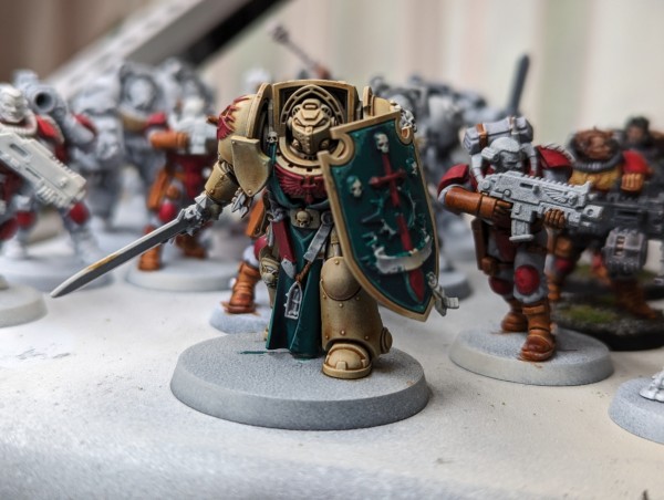 A partially painted Warhammer 40k miniature. A Dark Angels Deathwing Terminator in bone-colored armor with green cloth and shield.