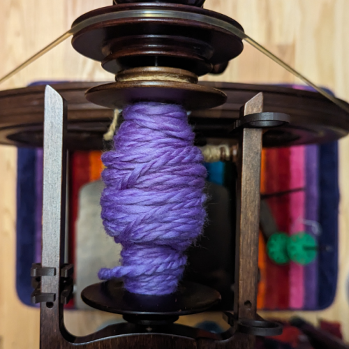 A bulky two ply purple yarn on a bobbin on a wooden spinning wheel. The view is top-down looking directly at the floor, where you can see that the wheel is sitting on a rainbow mat on a hardwood floor.