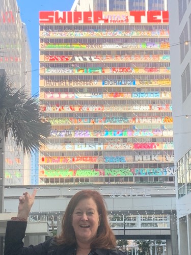 A woman smiles pointing up at a skyscraper building covered in graffiti. 