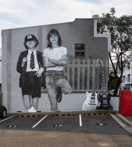 Streetartwall. The impressive, realistically designed mural with the portraits of the two rock musicians Angus and Malcolm Young (AC/DC) was sprayed/painted on the outside wall of a one-story gray building with a small window. The mural is in black and white and shows the two in their younger years. Angus with curly hair, in his typical short school uniform and baseball cap, stands casually next to his brother with half-length hair, white T-shirt and jeans. They are leaning against a painted fence. Next to them are two guitars, one white and one black. (The photo shows an empty parking lot in front of them and an old tree and a red clothes collection box on the right)