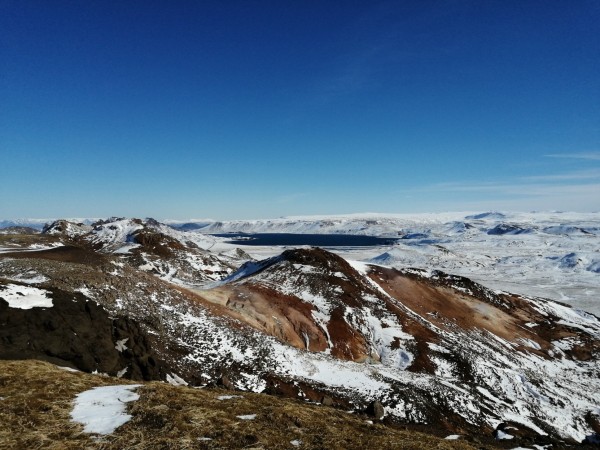 Une vue panoramique sur un paysage montagneux partiellement enneigé. La tâche bleu très foncé au fond indique la présence d'une grande étendue d'eau.