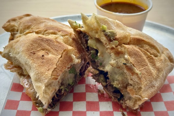 Mexican torta (with a side of Birria consomé) cut in half and stacked on a metal serving plate.