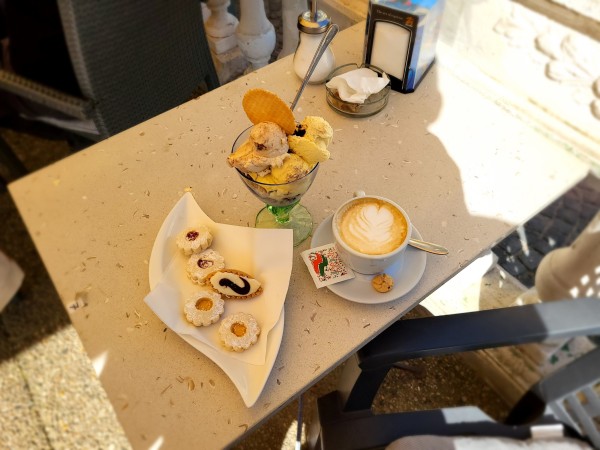 On the left a plate with cake, on the right a cappuccino, in the back a bowl with ice cream. Everything is on a table, and it's mostly in the shadow of a sunscreen.