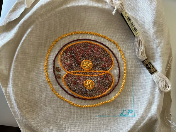 Two cells dividing embroidered on off white linen. A circle of brown stem stitch is the cell membrane, the corona radiata is in bright orangey yellow zig zag chain stitch. The edges of the dividing cells are in bright orange split stitch. The nucleolus of each cell is in bright yellow bullion stitch. The cytoplasm is stitched in variegated orange/brown thread in a mix of seed stitch and French knots. Some guide lines and Lia’s initials are marked in blue. A skein of off white embroidery thread sits on the edge. 