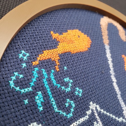 A close up of a cross stitch of an orange fish leaping.