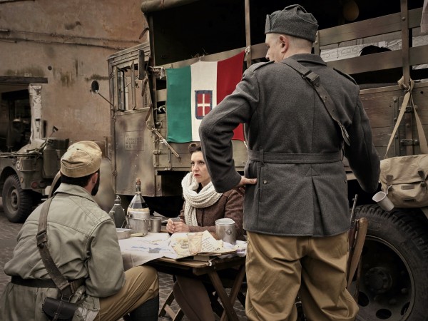 A modern photograph captures a poignant World War II reenactment scene in Italy, with two men and a woman gathered around a table with a map, planning or discussing strategies. The man facing away wears a grey military uniform, indicating his role, with a rifle slung over his shoulder, and a vintage Italian flag with the Savoy shield draped on the side, evoking the period's atmosphere. The setting includes a military vehicle and artifacts from the era, all contributing to a silent homage to the days of liberation.