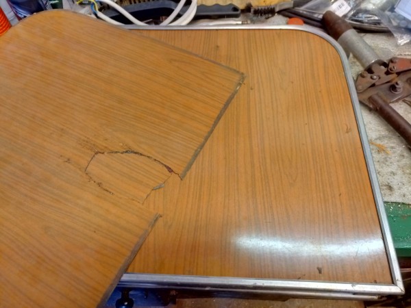 Old aluminium framed folding table, probably from Soviet times. A broken resin laminate top with fake wood patterning has been extracted already and rests on top of the table, a patched hole with a piece missing.