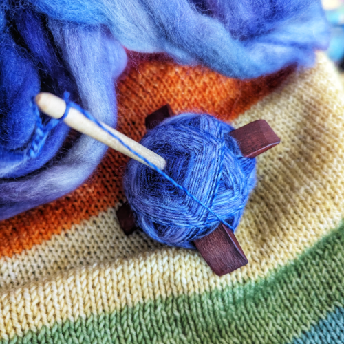 A purple/blue yarn in progress on a Turkish drop spindle. It is sitting on my kid's mostly finished rainbow sweater.