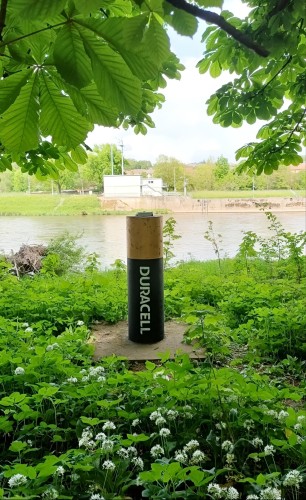 Streetart. A short, narrow concrete pillar on the riverbank has been transformed into a realistically designed 12-volt battery using spray paint. Instead of the ugly concrete, a black and gold Duracell battery now stands between trees and clover.
Info: The battery was designed back in 2021, but had become too weathered in the meantime. A redesign/recharge was urgently needed.