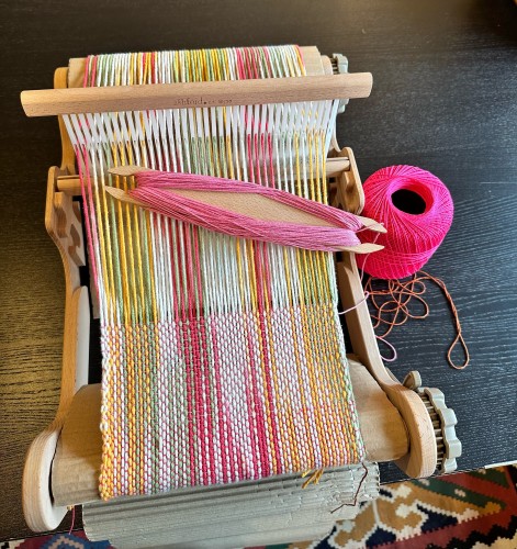 A loom is warped up with multicolored threads in yellow, green, and cream. A stick shuttle with pink weft sits on the loom. 