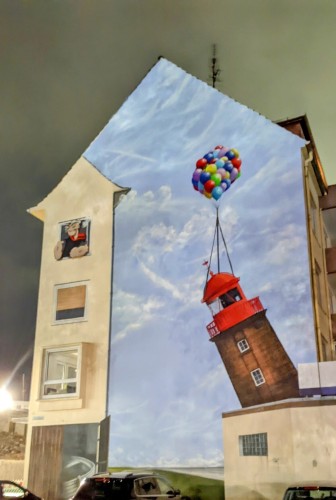 Zu sehen ist eine bemalte große Hauswand in Bremerhaven. Links oben schaut aus einem aufgemalten Fenster ein rauchender Popeye raus und rechts ist im Hintergrund Himmel mit Wolken. Davor ist ein gekippter Leuchtturm zu sehen, an dem oben eine große Menge zusammengebundener Luftballons befestigt sind, so dass sie scheinbar den Leuchtturm halten . Der Leuchtturm soll sicher den an der Nordmole darstellen, der 2022 durch die abgesackte Mole gekippt war und drohte deshalb umzukippen. Inzwischen wurde er abgetragen.