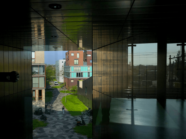 A series of reflective panels surrounding a view onto a living complex outside. Differnt buildings can be seen as well as a patch of green grass and a patchwork stone path.