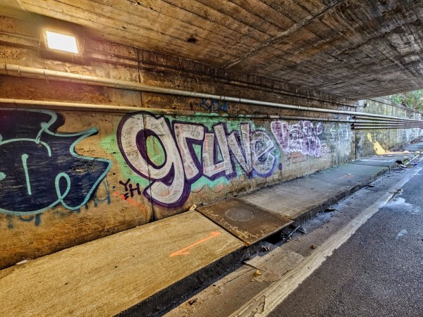 In a dark, dirty, weathered tunnel beneath an interstate highway bridge, the yellowish lighting illuminates colorful graffiti tagging the walls.