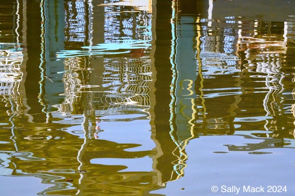Abstract color photo consisting of reflections in water of a docking pier. Most of the reflections are brown and turquoise verticals linked visually by smaller, thinner verticals, mostly white. Bright yellow reflections can be seen in the foremost turquoise veertical. The water is blue and brown. 