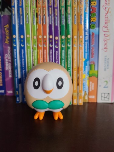 Photograph of a plastic model kit in front of books. The model kit is the Pokémon called Rowlet, which is a round, ball shaped light brown owl with a white face, green leaf-like shape on the center that looks like a bowtie. Behind the Rowlet figure are all of the volumes of the Pokémon Sun and Moon manga in order beginning with Volume 1 on the left. The Rowlet is looking head on toward the camera.