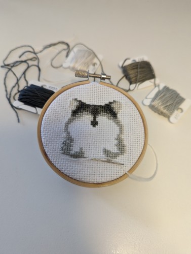 Picture shows an unfinished cross stitch project of a raccoon using different shades of grey threads.