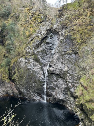 A narrow vertical stream of a waterfall flowing over dark rocks into a dark pool. 