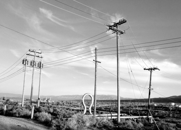 Black and white. Oil fields near Taft, .CA. Horizontal and vertical pipes and multiple electric poles rise from flat scrubby desert. 
Mountains are visible on the far horizon.