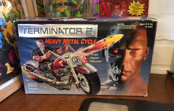 A desktop with on it a vintage boxed Kenner Terminator Heavy Metal Cycle toy. The artwork is amazing, popping, and hand-drawn. Cyborg steel and big fat bright letters everywhere.