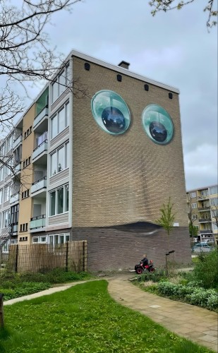 Streetartwall. A mural with a face was sprayed onto the exterior wall of a modern four-storey residential building. The beige-colored brick wall was decorated with two large black and white googly eyes, a long nose and an open mouth and looks very friendly. However, the house with a face was only on display for one day, as another large mural is being created here. A bit of fun, because the artist has already painted over it again and is now redesigning the wall in large areas of color. 12 Apr. 2024. 
(The photo shows a little boy on a small red quad bike in front of the mural, with green bushes, a view of other buildings on the right and a straw fence in front of the façade on the left)