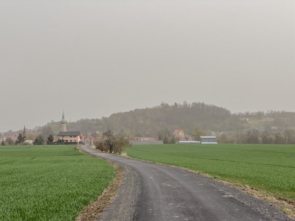 Pochmurny wiejski krajobraz z zielonym polem, krętą drogą na pierwszym planie i wioską z iglicą kościoła w oddali.