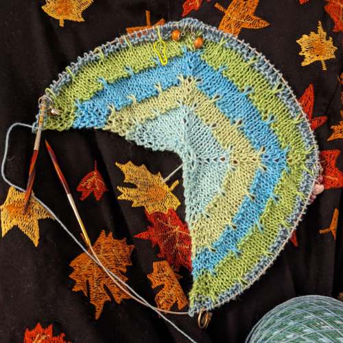 A shawl in progress using self striping shawl yarn. This first section is all light blues and greens which contrasts with the embroidered fall leaves on my black dress. 
