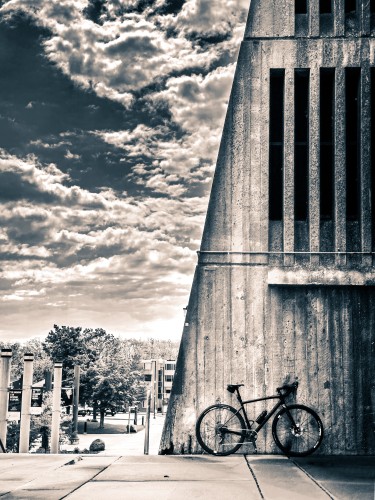 monochrome photo of a black specialized diverge gravel bike standing against an angled concrete wall. The concrete wall has dark slits in it and looks like it’s reaching up to the sky. To the left of the wall is a dramatic sky, full of puffy clouds. 